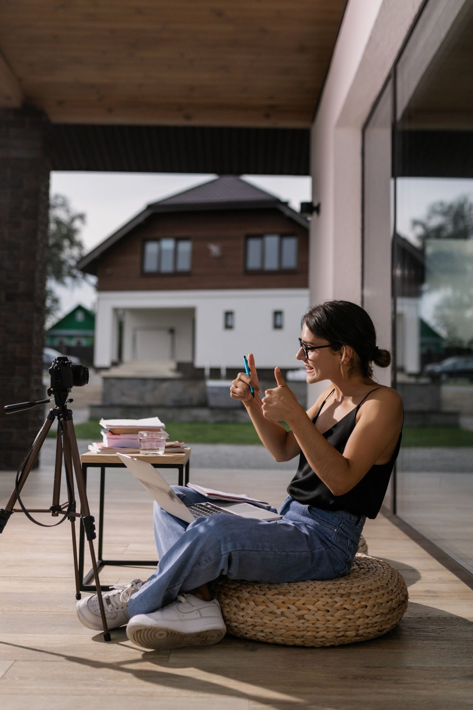 Young woman with eyeglasses posing with camera and laptop, showing thumbs up. Casual summer vibe.
